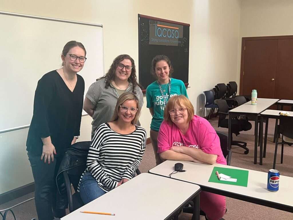 Three library employees pose smiling with two adult students at a computer class.