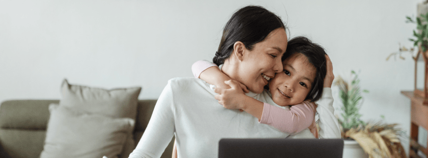 mom turning toward daughter who is hugging her from behind as she works on her laptop
