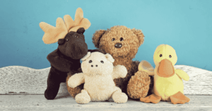Four stuffed animals (a moose, pig, teddy bear, & duck) sit on a white wooden shelf, in front of a blue wall.