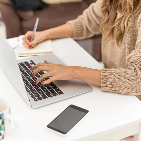 woman typing on a laptop with one hand and taking notes with the other